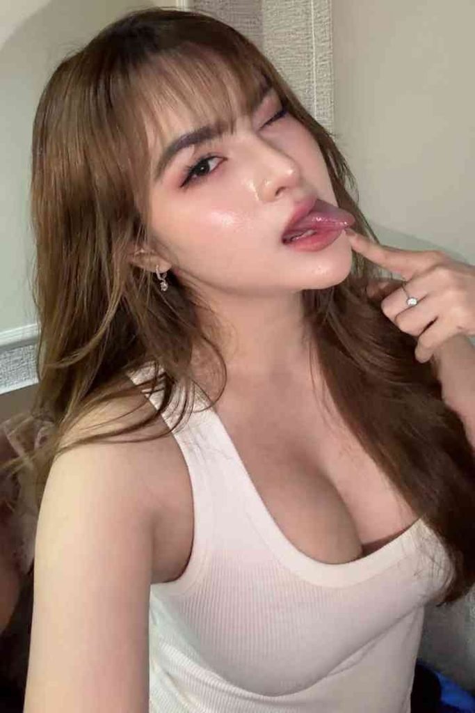 KL Escort Girl Young woman with long brown hair and bangs, wearing a white tank top, winks and sticks out her tongue. Malay Escort